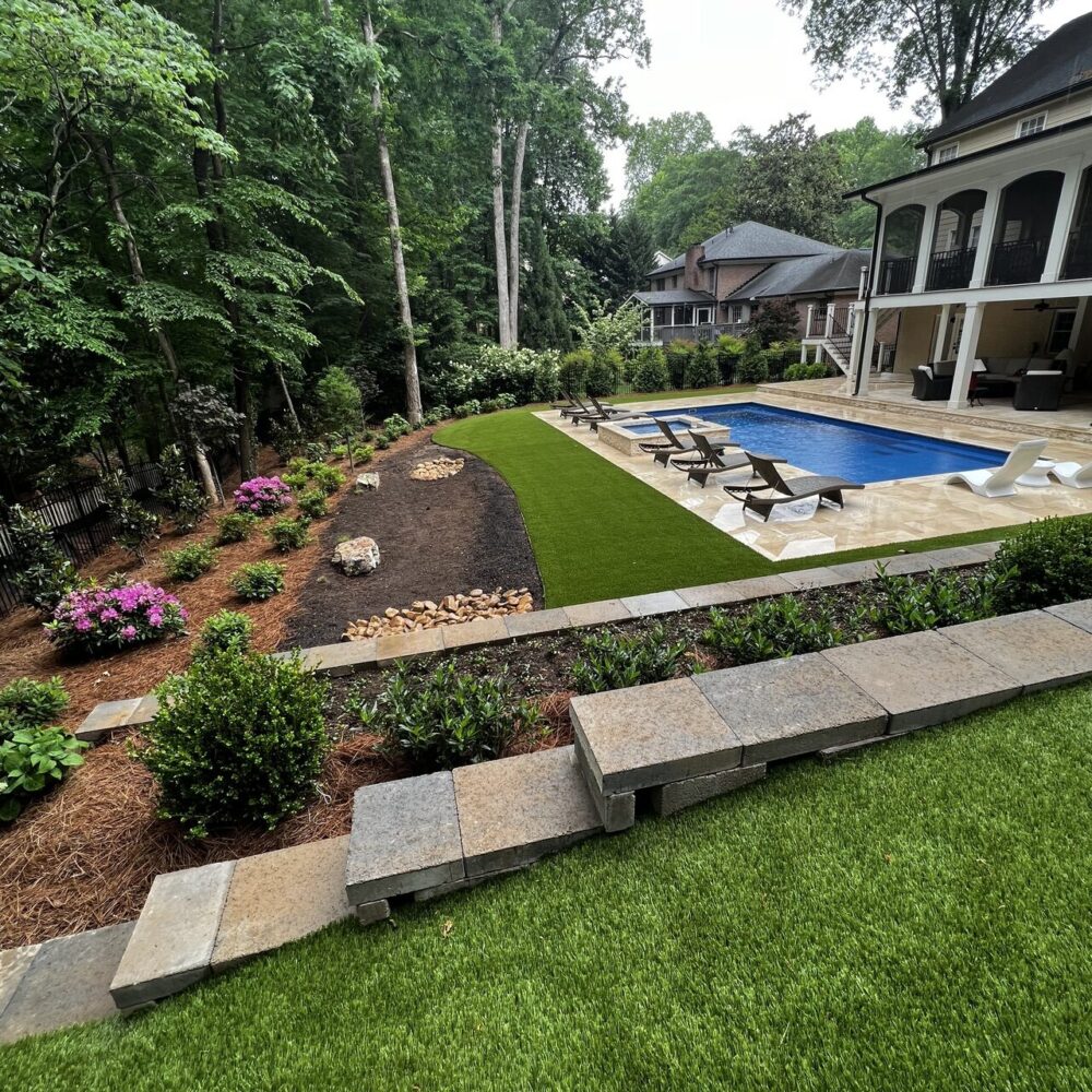 Completed backyard renovation featuring a swimming pool with a travertine deck, surrounded by artificial turf. The area is enhanced with various landscaping elements and bordered by retaining walls.