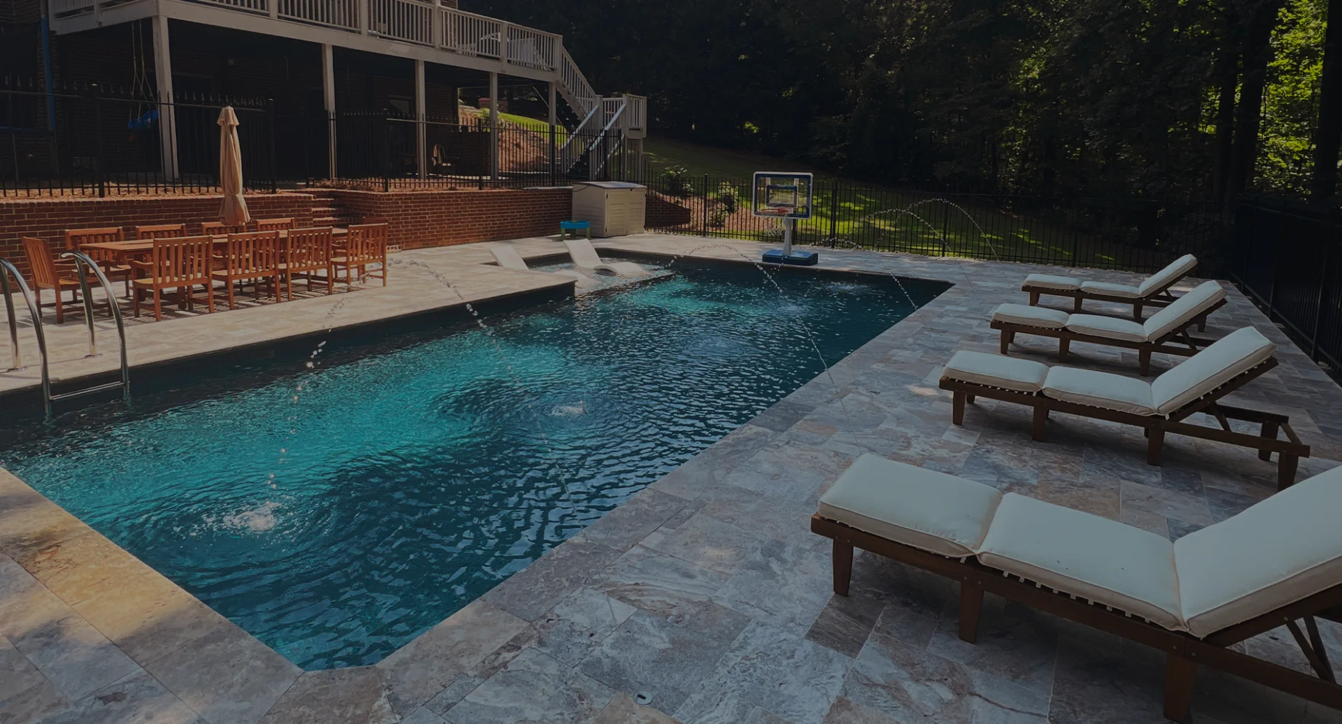 Free-form fiberglass pool surrounded by ivory travertine stone, a raised lounging area that includes two water features, and LED lights illuminating the inside of the fiberglass pool.